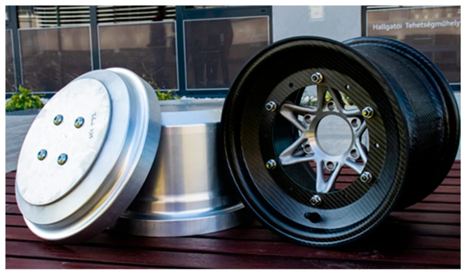 A silver aluminum disc-shaped laminating tool beside a finished three-piece CFRP rim with a star-spoked 3D-printed center and glossy woven-carbon barrel.